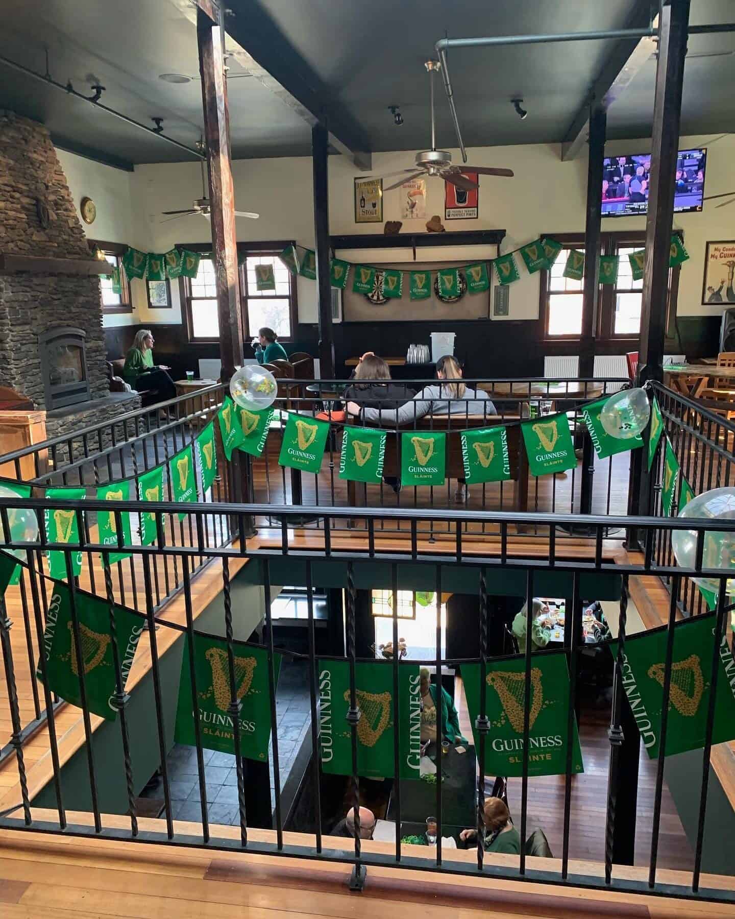 A pub interior decorated for St. Patrick's Day, featuring Guinness banners, a stone fireplace, a staircase, and patrons seated on both levels.