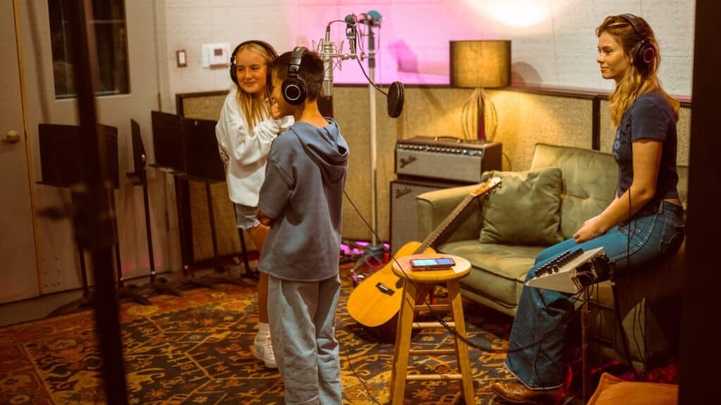 Three people wearing headphones are in a recording studio; two stand by a microphone while one sits on a stool near a guitar and audio equipment.