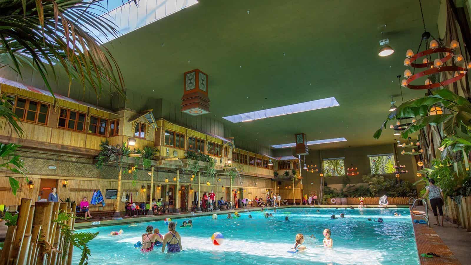 Indoor pool in Bothell with children and adults swimming and playing with beach balls. The area features high ceilings, skylights, and tropical decorations. Several people are seated around the pool's perimeter, enjoying this hidden Washington gem.