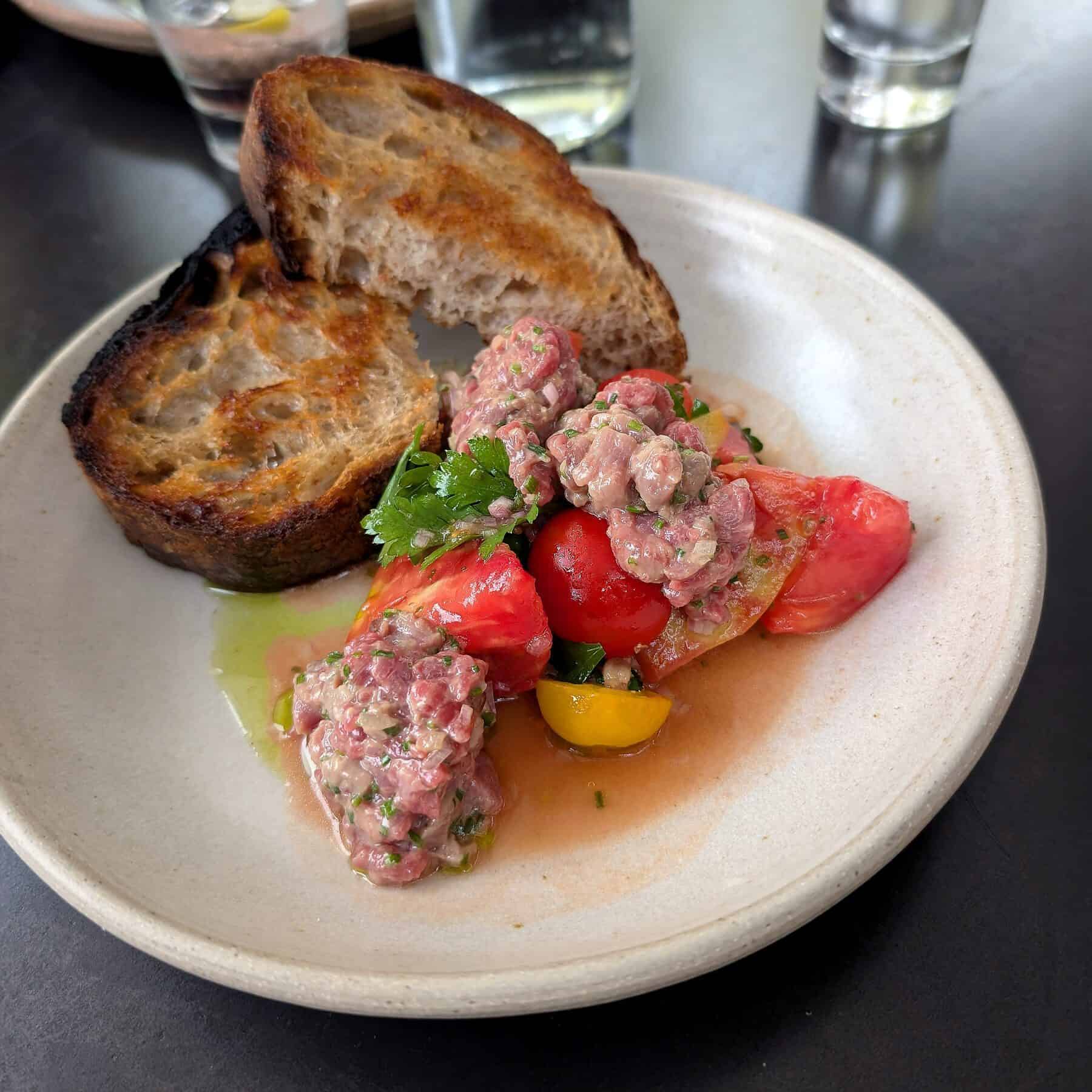 Plate with toasted bread slices, chopped beef tartare mixed with herbs, and assorted tomato wedges—an ideal choice among seasonal foods—served on a white dish with a glass of water in the background.