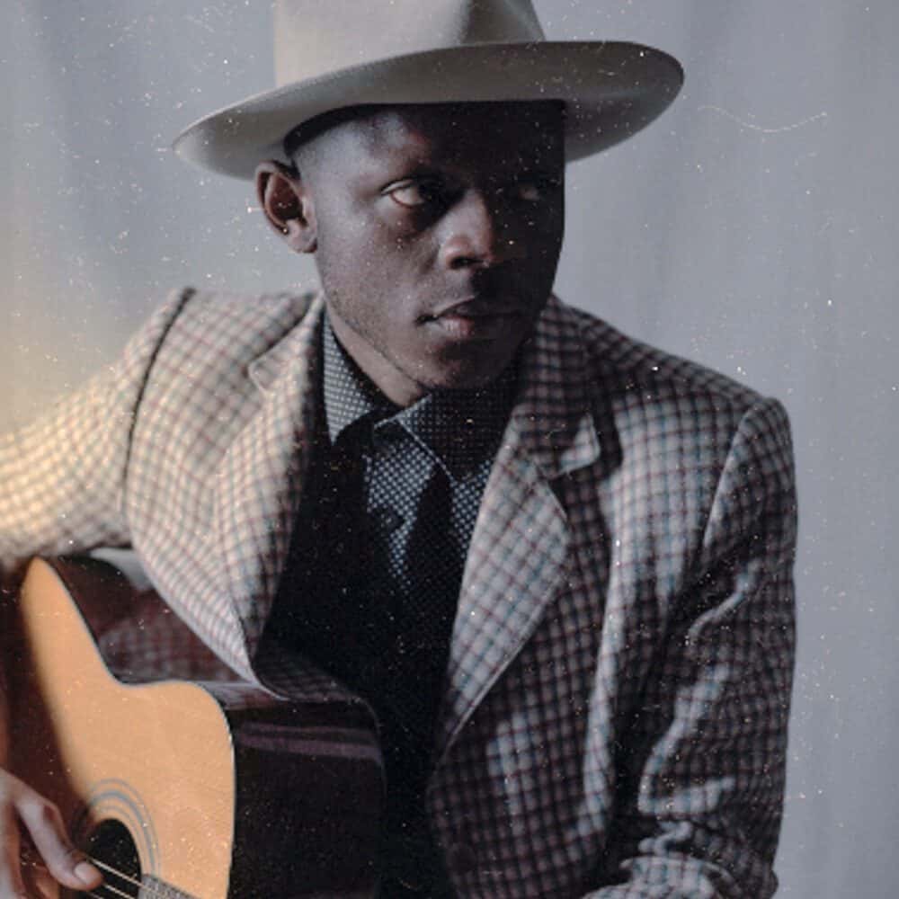 A man wearing a plaid suit, polka dot shirt, and wide-brimmed hat sits holding an acoustic guitar, looking slightly to his left against a neutral background—a Must List highlight for 09-04-2025.