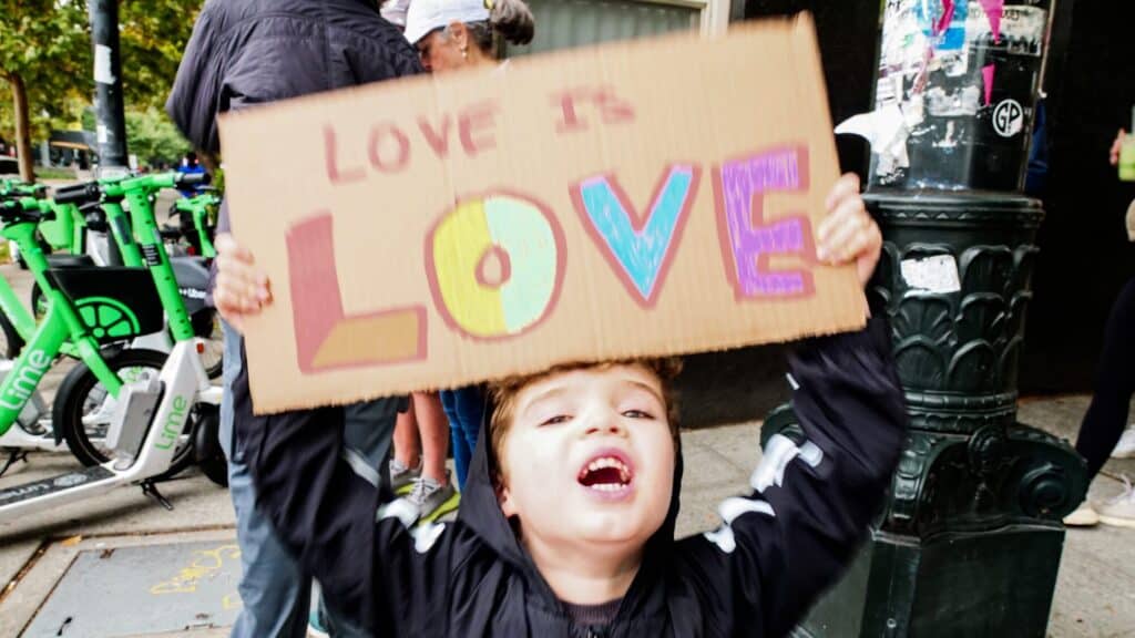 Young child holding up a cardboard sign that reads "LOVE IS LOVE" with brightly colored letters on a city sidewalk.