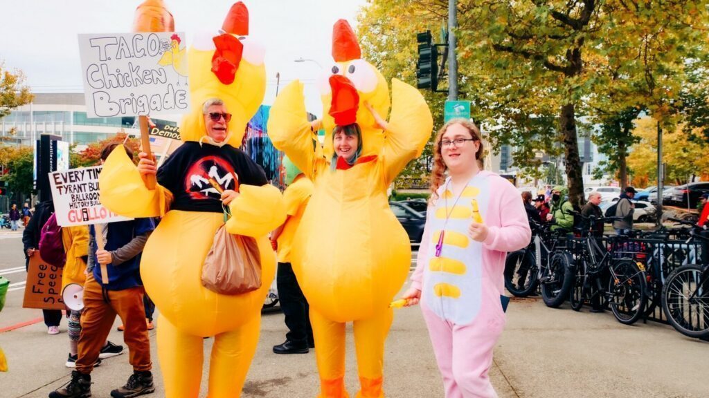 Three people in costume stand outside: two in inflatable chicken suits and one in a pink onesie. They are holding protest signs. Bicycles and trees are visible in the background.