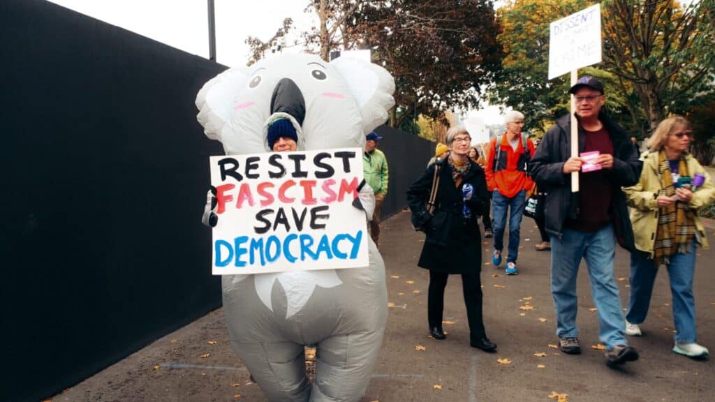 A person in a koala costume holds a "Resist Fascism, Save Democracy" sign while walking in a protest with other people carrying signs.
