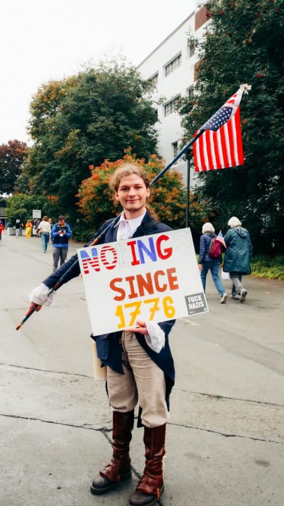 Person in historical costume holds an American flag and a sign reading "NO KINGS SINCE 1776" at an outdoor event with other people and trees in the background.