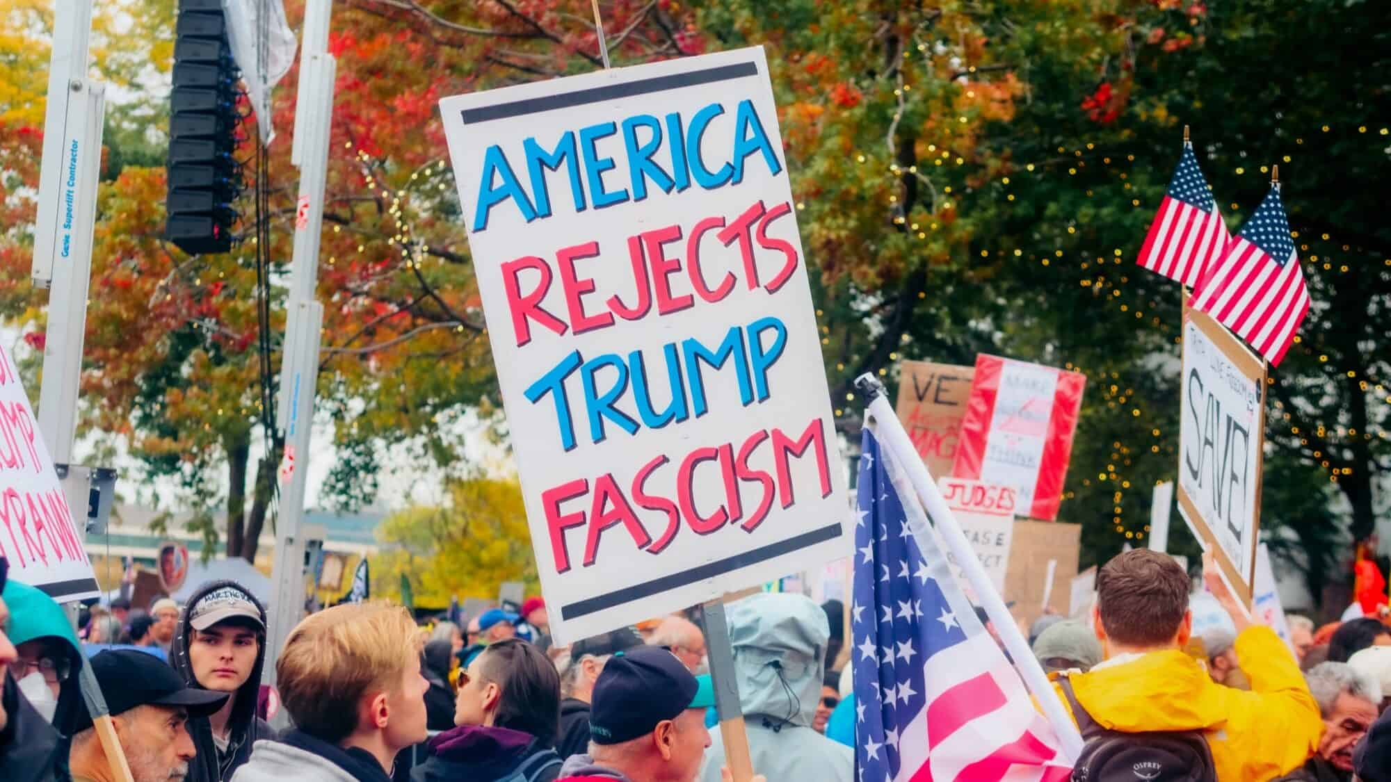 A crowd of people at a protest holds signs, including one that reads "America Rejects Trump Fascism," and several American flags are visible.