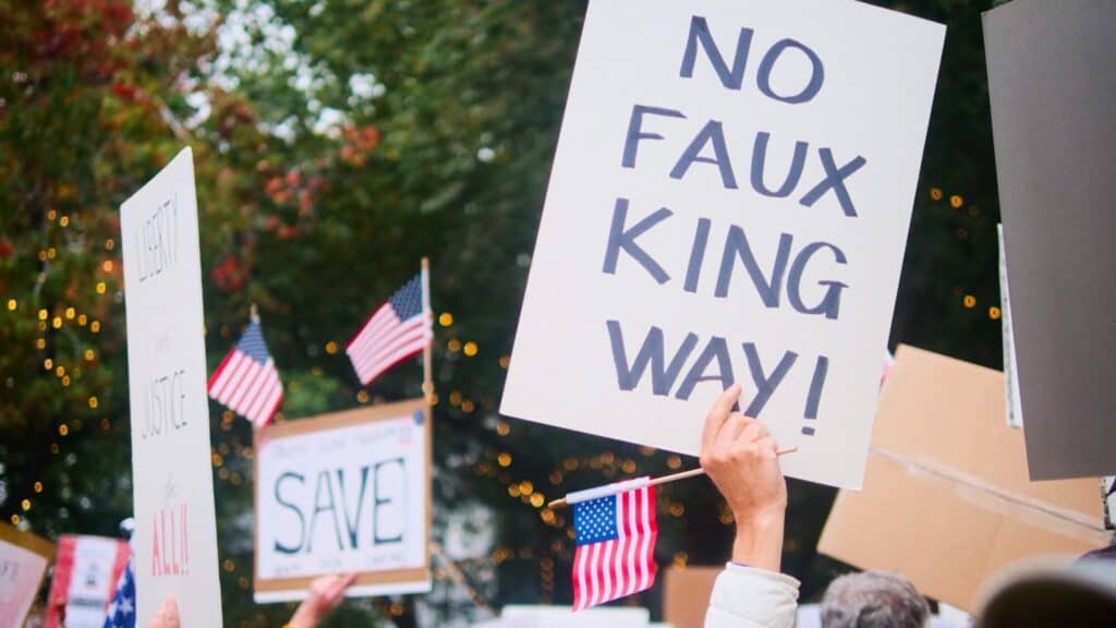 A protester holds a sign that reads "NO FAUX KING WAY!" among a crowd with American flags and other signs. Trees and lights are visible in the background.