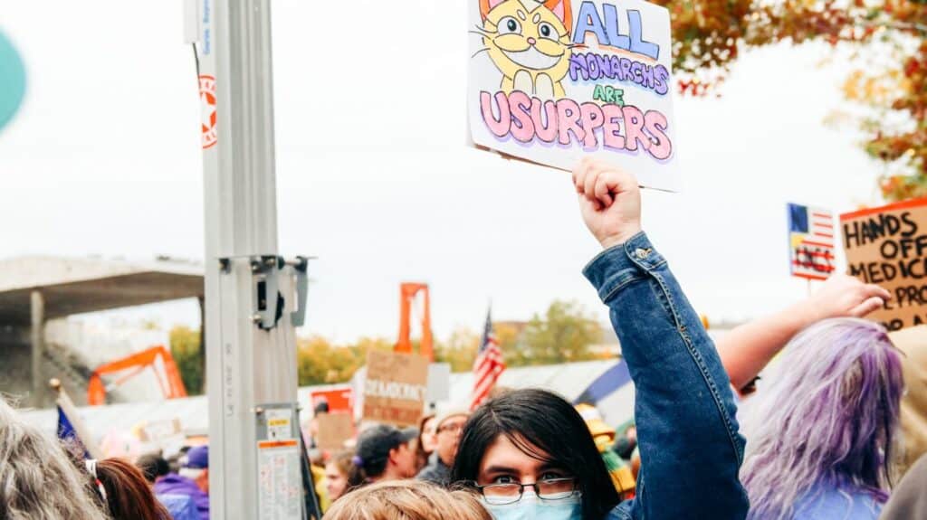 Person in a crowd holds a colorful protest sign with a cartoon cat and the words "ALL MONARCHS ARE USURPERS." Other protest signs and people are visible in the background.