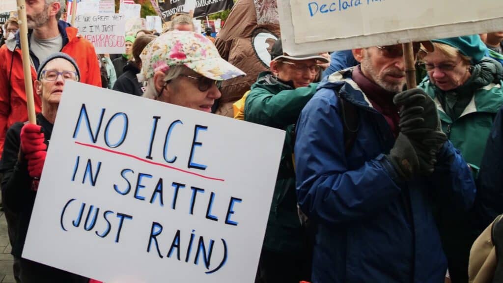 A group of people at a protest. One person holds a sign that reads, "NO ICE IN SEATTLE (JUST RAIN)." Other protest signs are visible in the background.