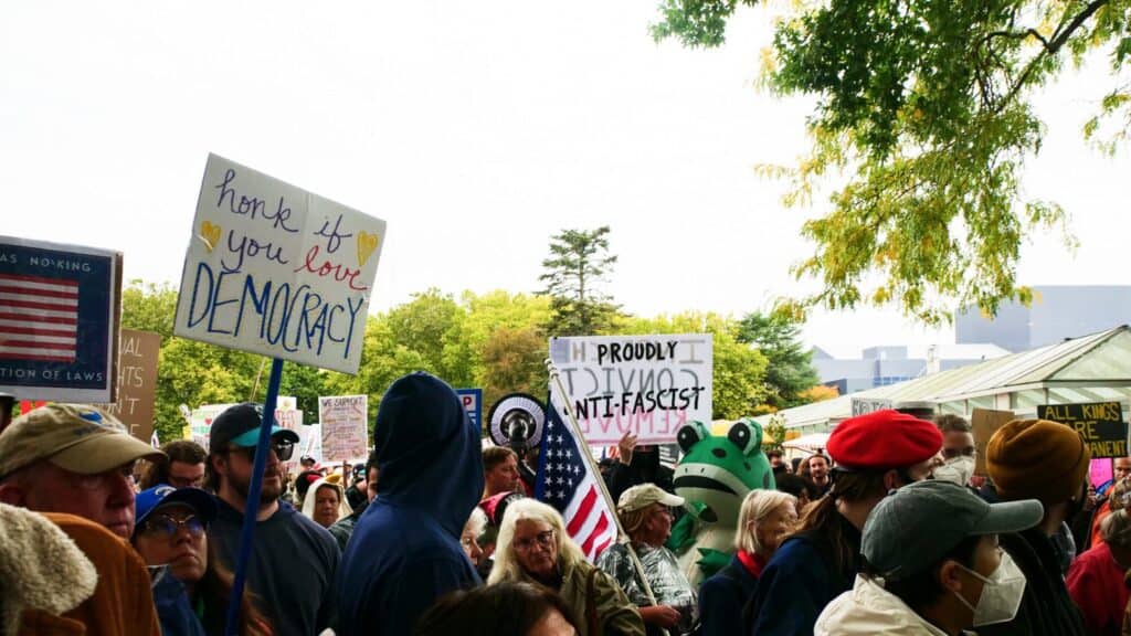 A large crowd of people at a protest holds various signs, including ones that read "honk if you love DEMOCRACY" and "PROUDLY DIVORCED ANTI-FASCIST.