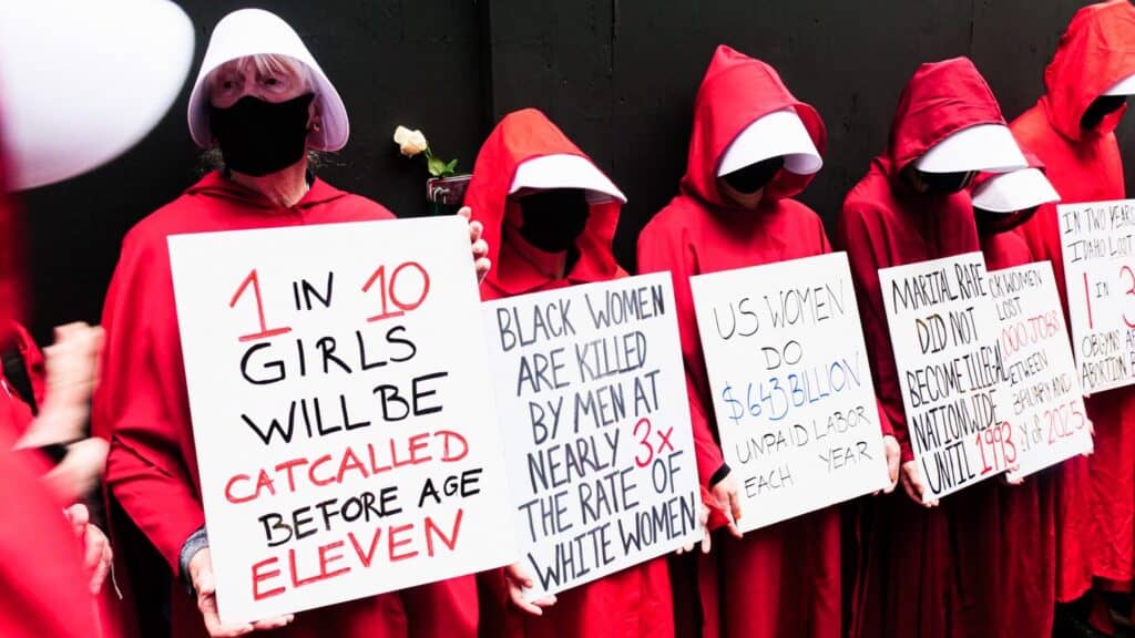 A group of people in red cloaks and white bonnets hold protest signs displaying statistics about violence and discrimination against women and girls.