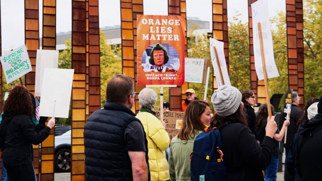 A group of people at a protest hold signs, including one reading "Orange Lies Matter" with a cartoon and others with various messages, standing in front of tall orange structures.