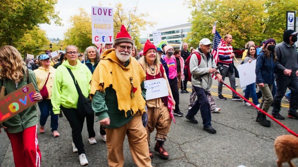 A group of people march outdoors, some holding signs and flags. Two people in fantasy costumes walk in front, surrounded by others in casual clothing. Trees and buildings are visible in the background.