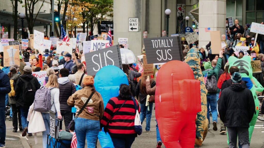 A crowd of protesters holding signs, including two people in inflatable crewmate costumes from "Among Us" with signs reading "SUS" and "TOTALLY SUS.