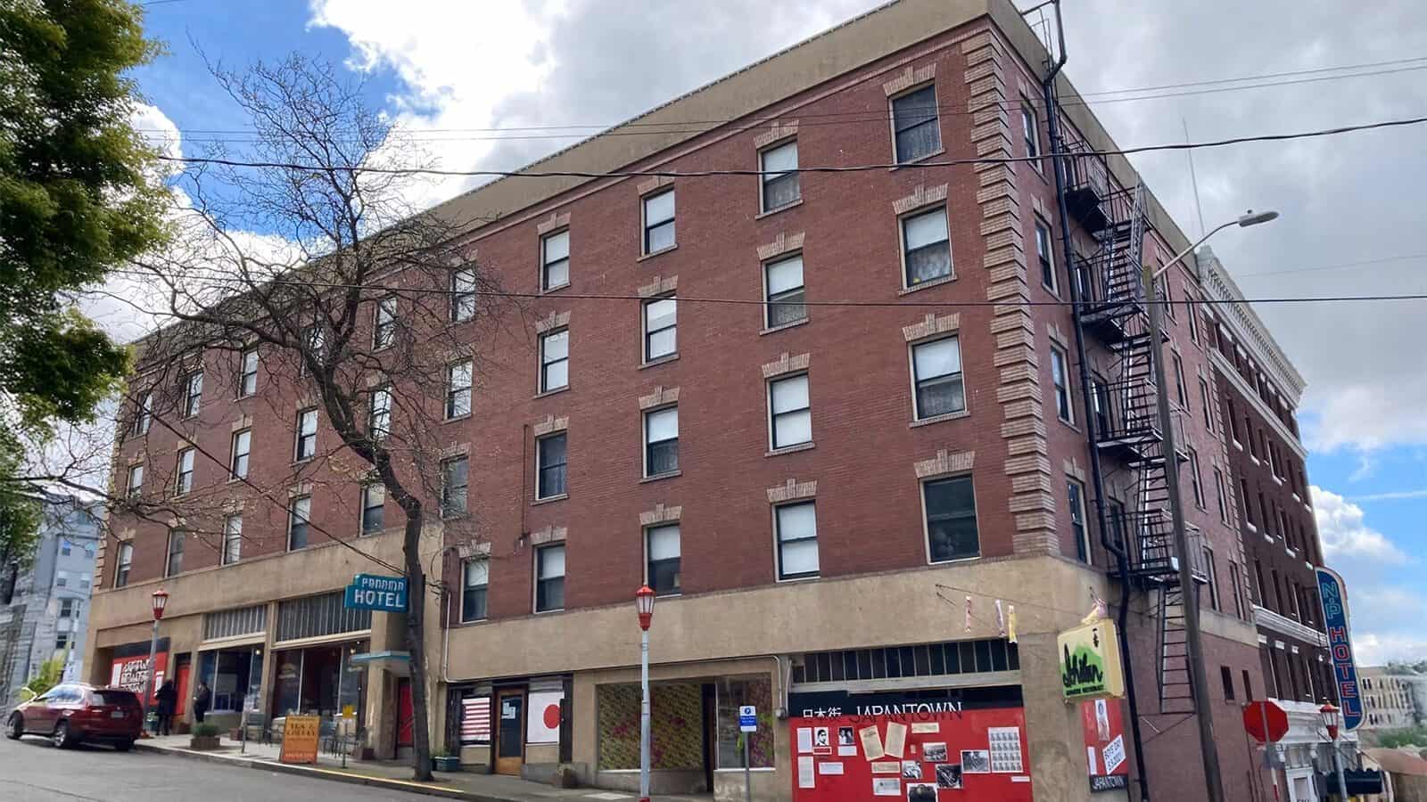 A four-story red brick building with a vintage hotel sign, fire escape stairs, and various posters on the lower windows, this Seattle landmark sits on a sloped street and reflects the city’s rich cultural heritage.