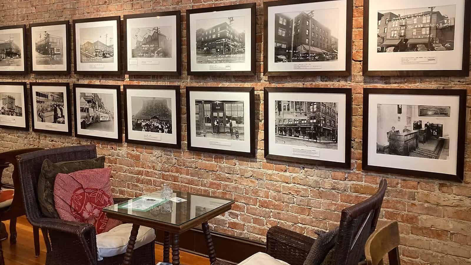 A brick wall displays framed black and white historic photos of Seattle heritage above a small glass table with wicker chairs and cushions in a cozy indoor seating area.