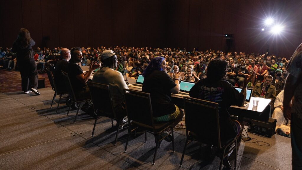 A panel of speakers sits at a table facing a large audience in a dimly lit conference room, with a bright light shining behind them.