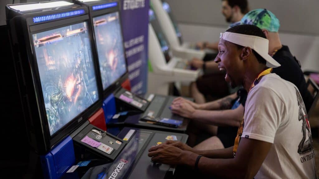 A man enthusiastically plays an arcade fighting game on a gaming machine, with other players seated beside him, at a gaming event.