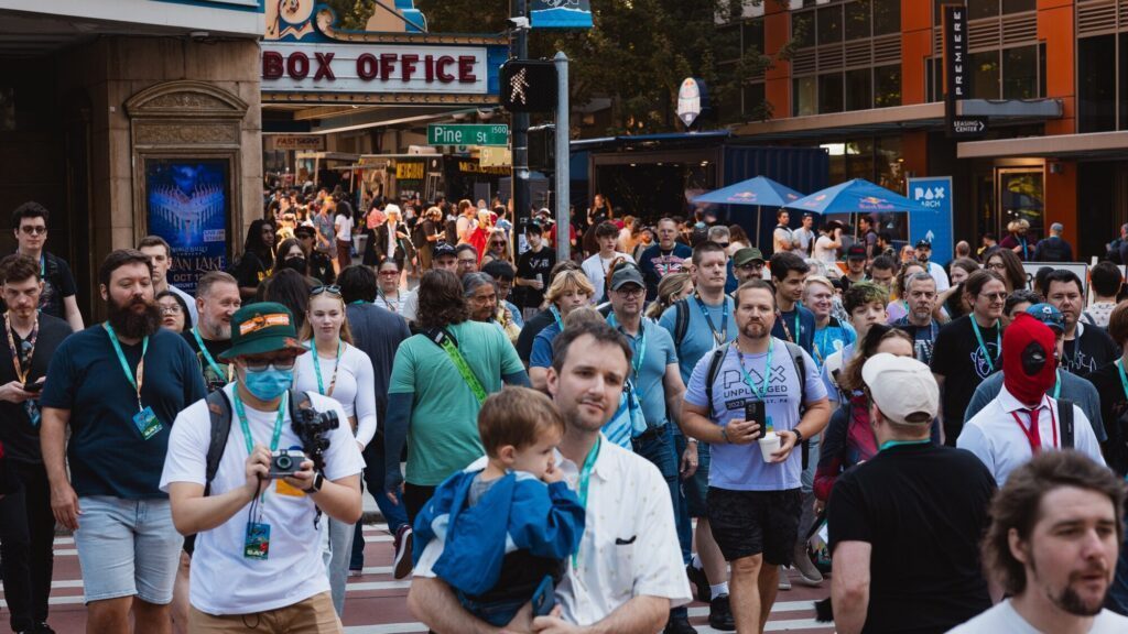 A large crowd of people, some wearing badges and cosplay, cross a city street near a box office and event signs on a sunny day.