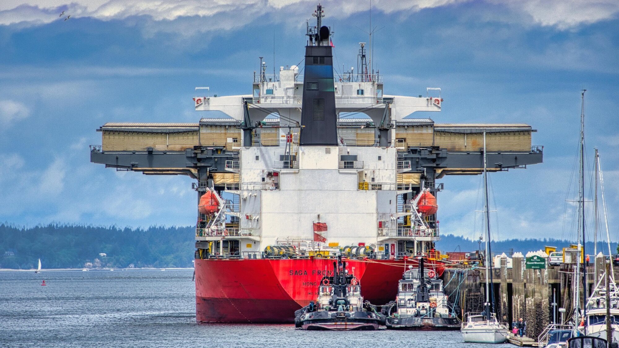A large red cargo ship docked at a port, with several smaller boats and tugboats alongside, and cloudy skies in the background.