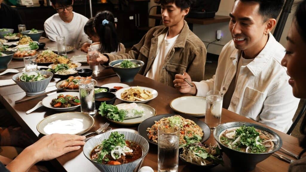 A group of people sit around a dining table filled with various Asian dishes, including noodles, salads, and greens, sharing a meal together.
