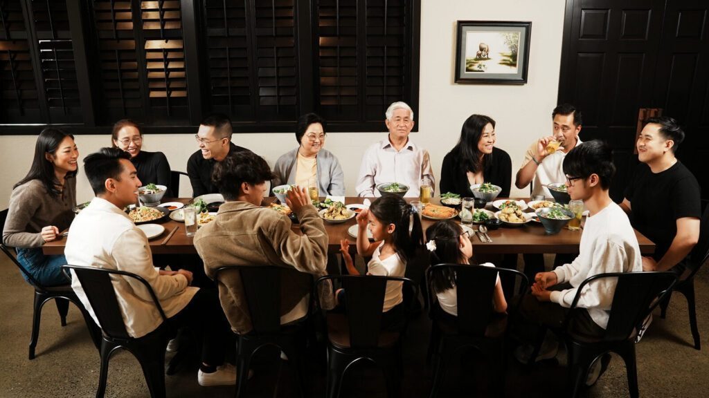 A large family gathers around a dining table, sharing a meal and engaging in conversation with bowls and plates of food in front of them.