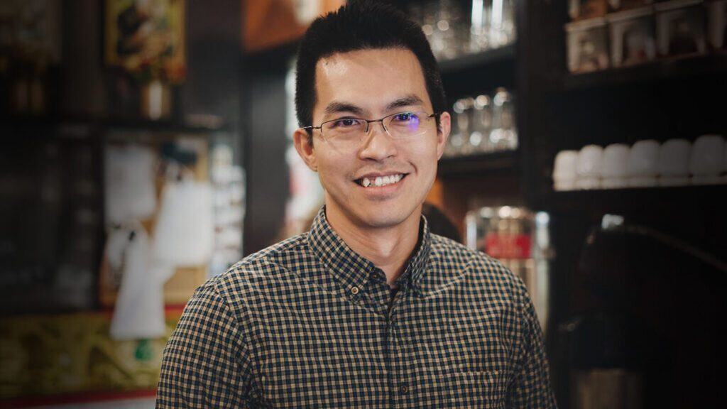 A man wearing glasses and a checkered shirt smiles while standing indoors, with shelves and glassware visible in the background.