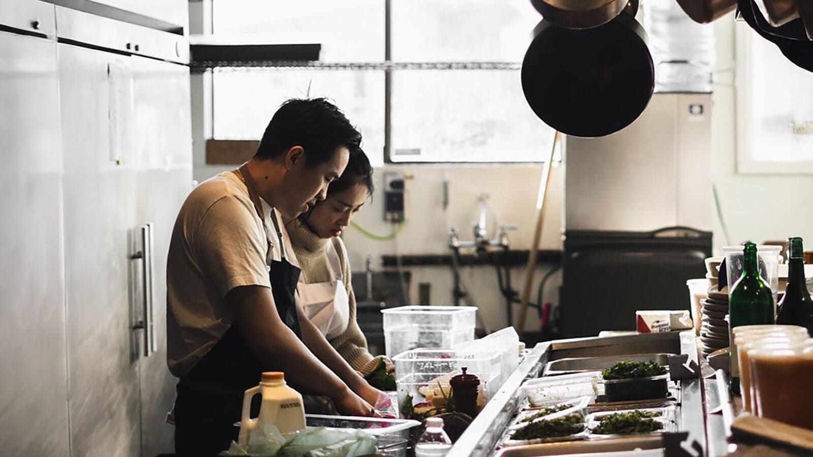 Two people are preparing food at a kitchen counter, surrounded by various cooking supplies and ingredients.