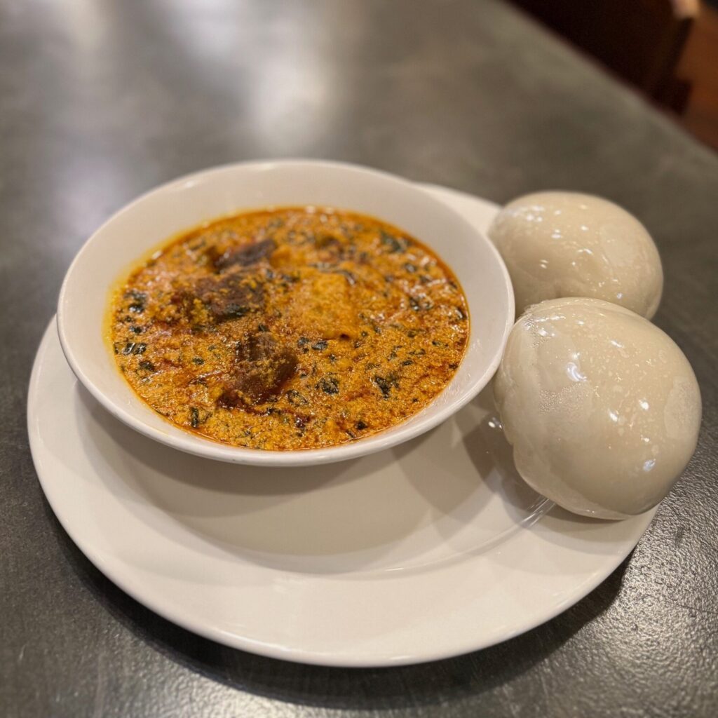 A bowl of egusi soup with pieces of meat, served on a white plate alongside two servings of pounded yam.