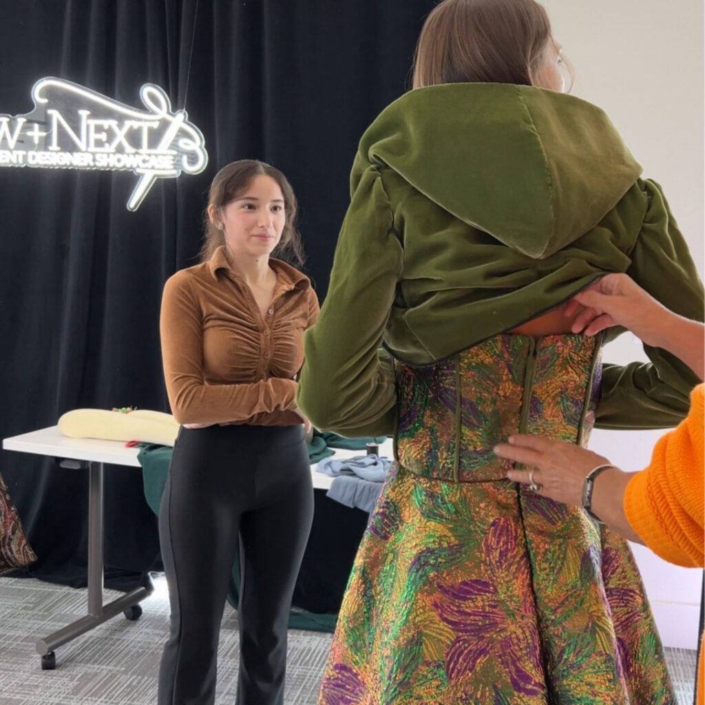 A woman in a brown shirt observes as another woman in a green jacket and colorful dress is being fitted by a person in an orange sweater at a fashion event.