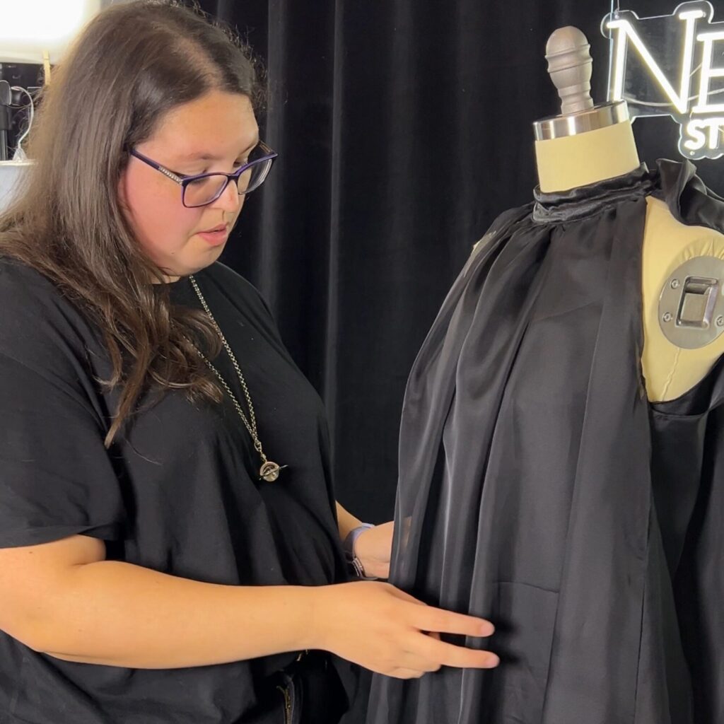 A person adjusts a black satin garment on a dress form in a studio with black curtains and a lit sign in the background.