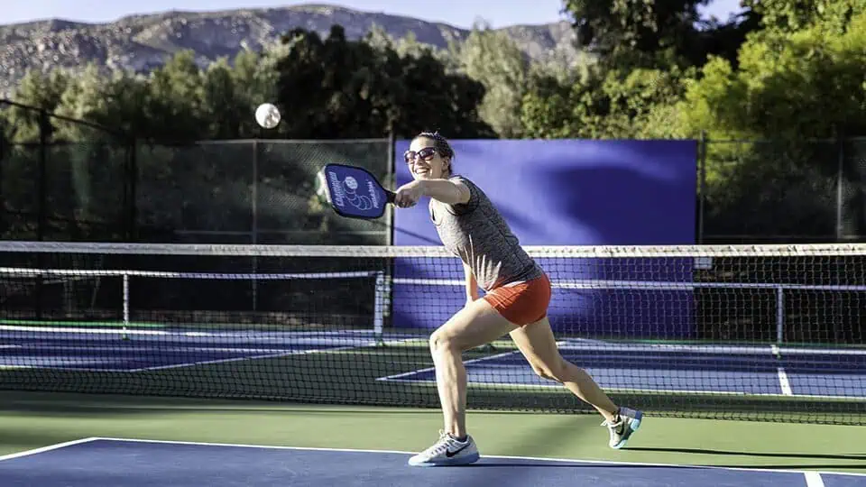 A woman in athletic clothing reaches forward to hit a pickleball with a paddle on an outdoor court at Rancho La Puerta, with trees and mountains creating a natural sanctuary in the background.