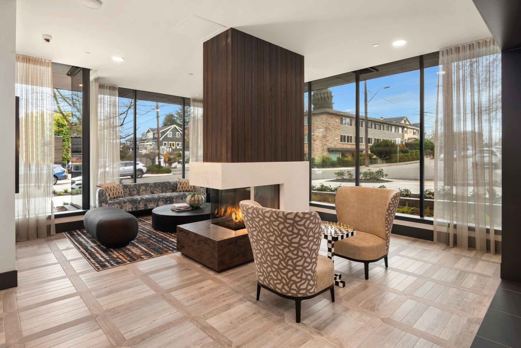 A modern seating area features cozy chairs and a chess table by the central fireplace, all beneath elegant Queen Anne gables. Surrounding large windows offer views of residential buildings and the street outside.