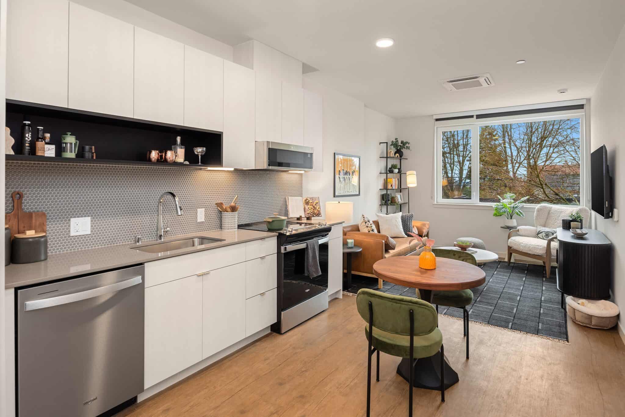 A modern open-plan kitchen and living area featuring elegant white cabinetry, a round dining table, and a cozy seating spot with a large window beneath stylish gables, all complemented by light wood flooring.