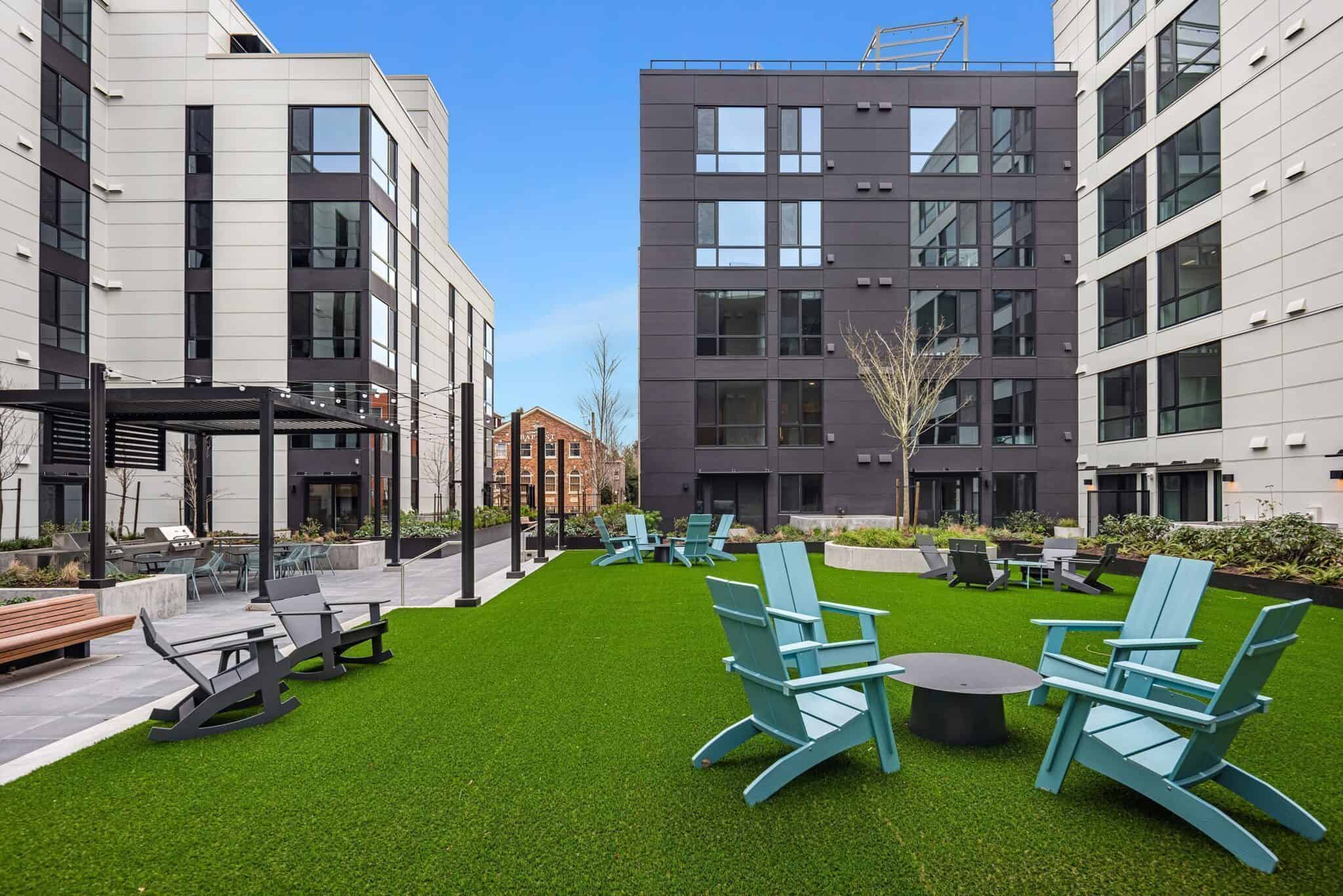 This modern apartment courtyard, reminiscent of the Queen Anne style, features artificial grass, Adirondack chairs, and inviting fire pits nestled among elegant Gables-style buildings.