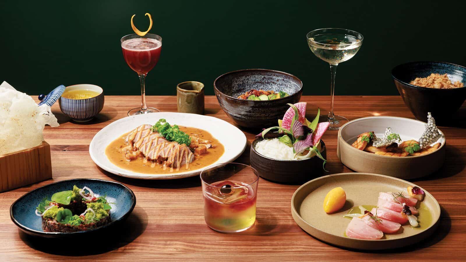 A wooden table set with various plated Vietnamese cuisine, gourmet dishes by Trinh Nguyen, cocktails, and a cup of tea, presented in a fine dining style against a dark green background.