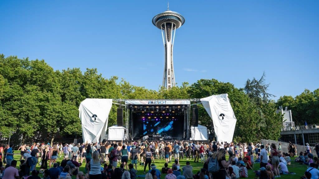 A crowd gathers on a lawn in front of a music stage with the Space Needle visible in the background under a clear blue sky.