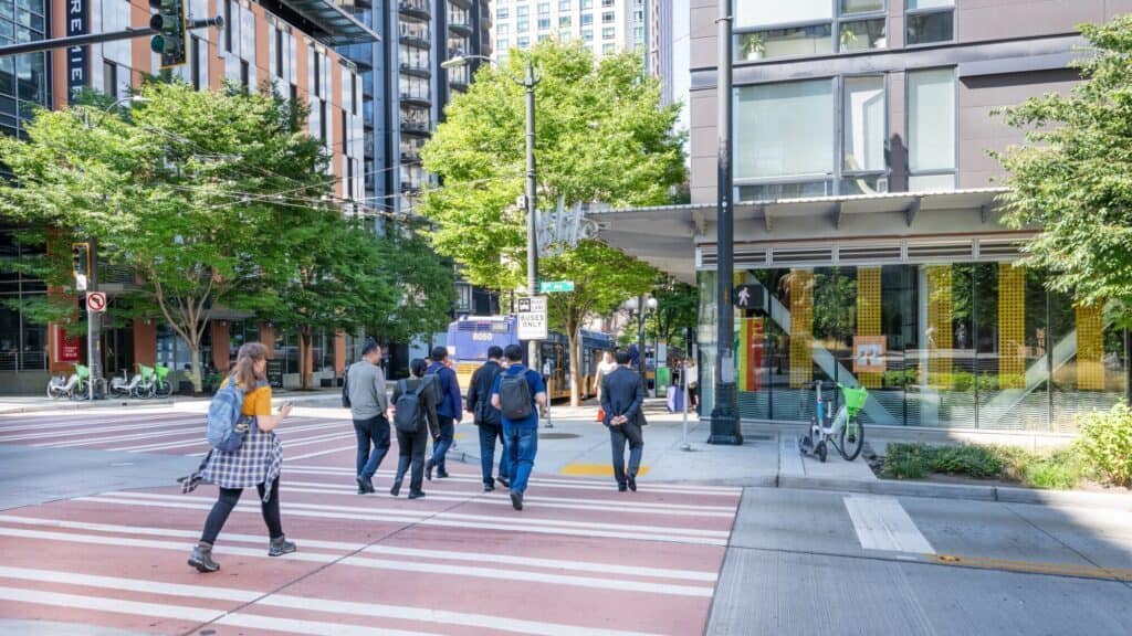 Several people walk across a city crosswalk on a sunny day, surrounded by modern buildings, trees, bike racks, and parked bicycles.