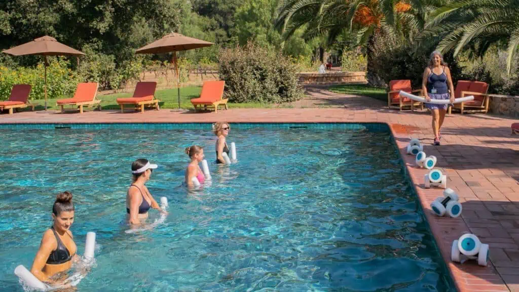 Four women stand in a swimming pool holding foam dumbbells as an instructor at Rancho La Puerta guides them. Red lounge chairs and umbrellas create a sanctuary-like atmosphere in the background.