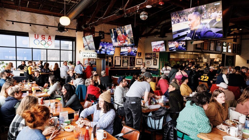 A crowded sports bar with people eating at tables, servers working, and multiple TVs displaying various sports events. USA Olympics banner hangs on the wall.