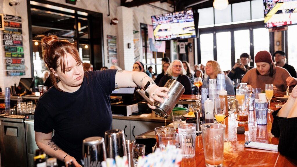 Bartender pours a drink behind a busy bar, with patrons seated at the counter and tables, and sports games displayed on TV screens in the background.