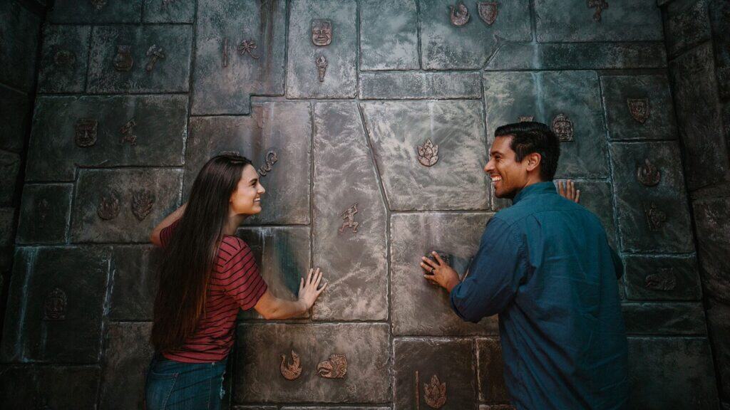 Two people stand facing a stone wall with engraved symbols, each pressing their hands against different sections and smiling at each other.