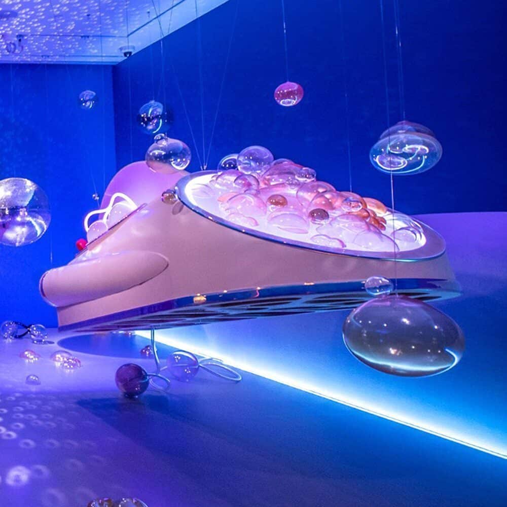 A large, futuristic installation shaped like a stylized animal head with illuminated details stands as a Must List highlight, surrounded by floating translucent bubbles in a blue-lit room—experience it before 08-28-2025.