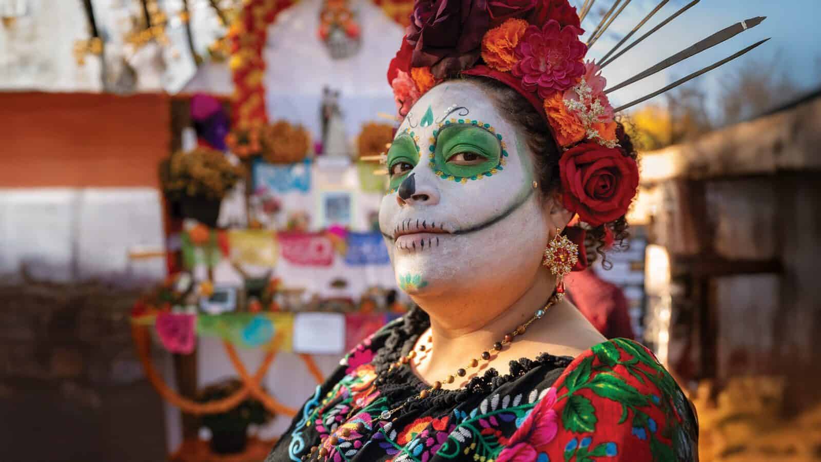 A woman dressed in traditional clothing with Day of the Dead face paint stands in front of an altar decorated with flowers and offerings, celebrating in the heart of Santa Fe, the vibrant City Different.