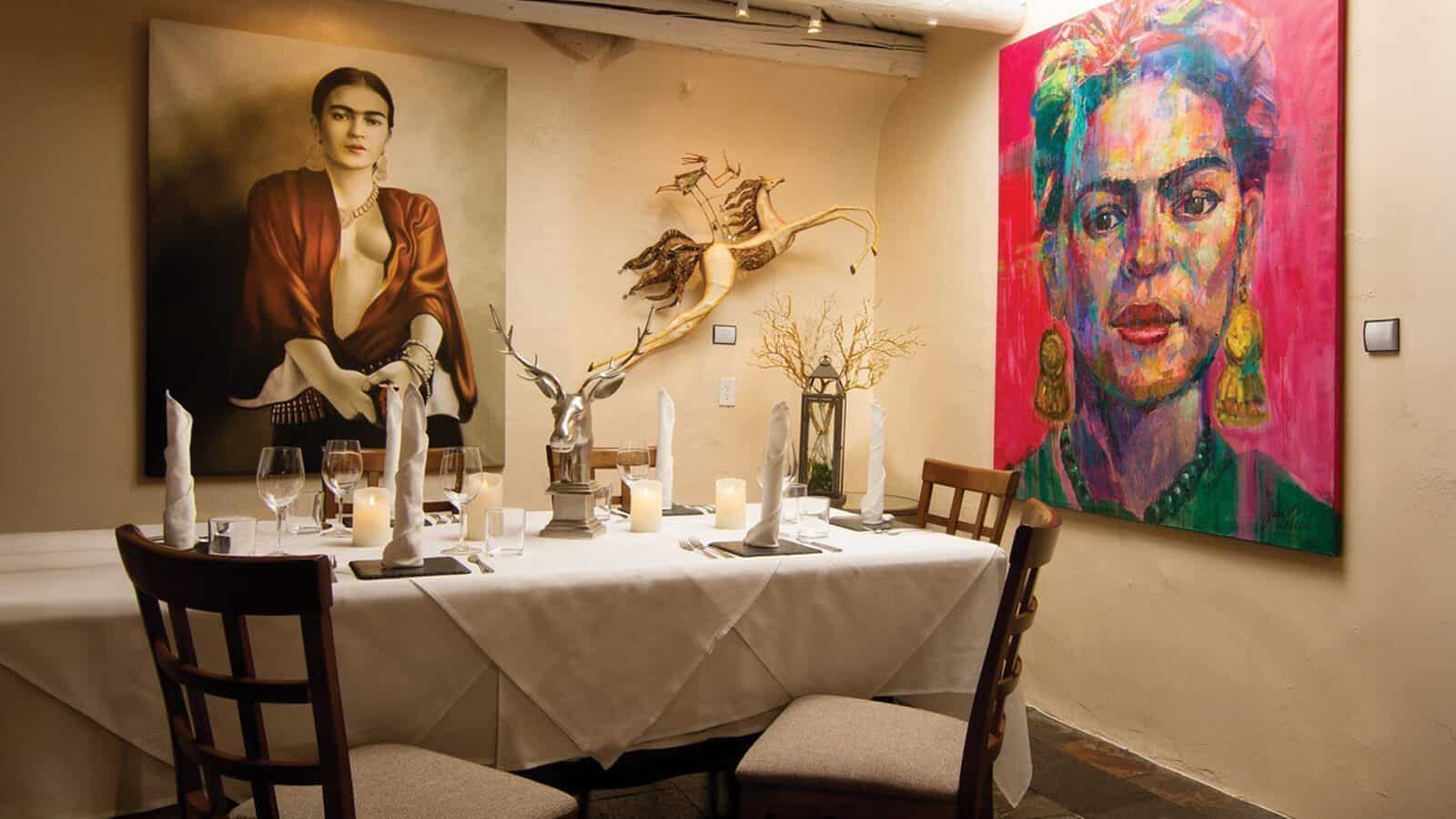 A dining table set for four sits between two large portraits of Frida Kahlo—one realistic, one colorful—with antler decor and candles, capturing the vibrant spirit of the City Different, a unique southwestern city influenced by Pueblo tribes.