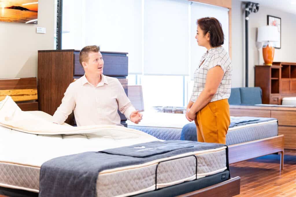 A man seated on a mattress in the furniture store chats with a standing woman, both envisioning more spacious bedrooms.