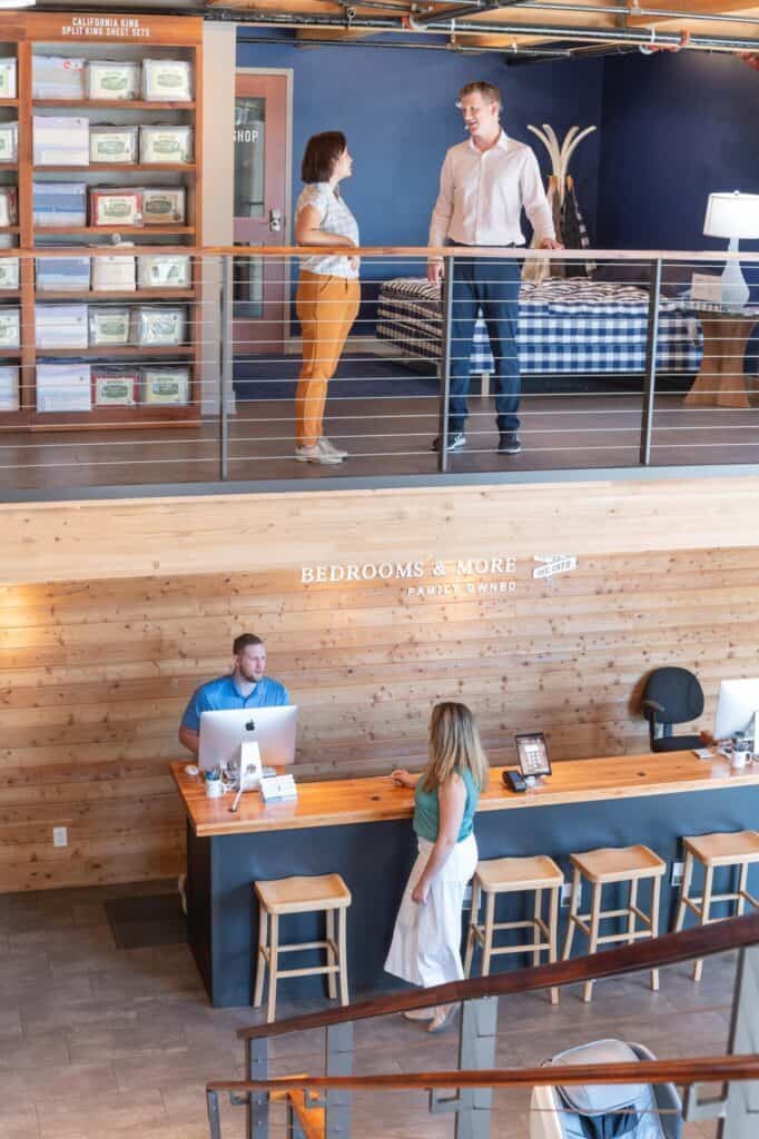 Two people stand on a mezzanine talking. Below them, a person sits at a desk with a computer while another stands by the counter under signs that read "BEDROOMS & MORE" and "FAMILY OWNED," capturing the essence of home comfort and more than just furniture shopping.