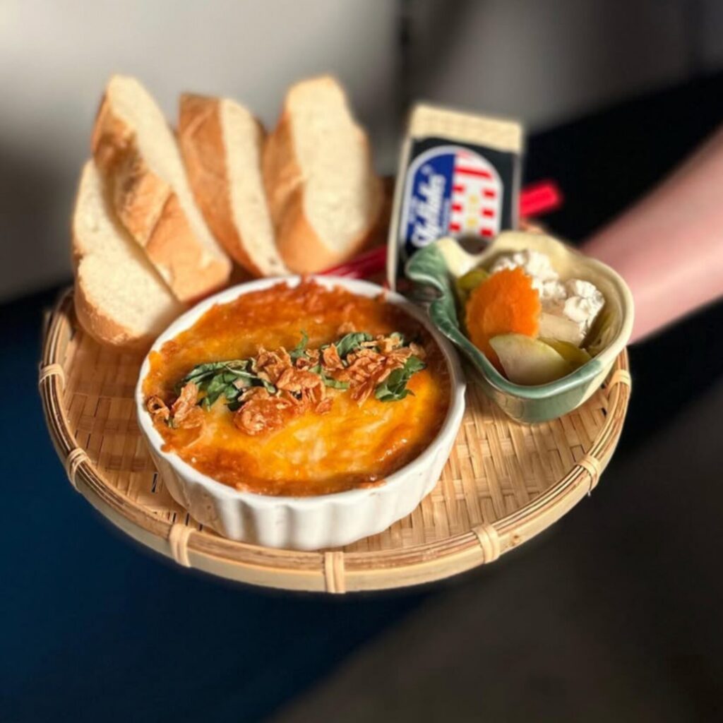A round tray with a baked dish topped with herbs, four slices of bread, a small bowl of pickled vegetables, and a pack of processed cheese.