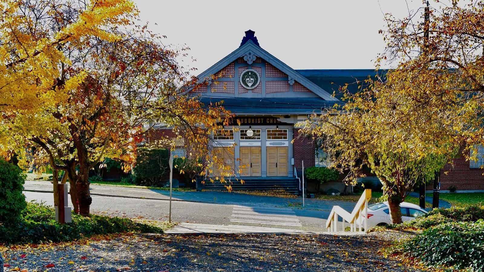 A brick building with a gabled roof and clock above the entrance is surrounded by autumn trees in vibrant yellow and orange. This Seattle cultural landmark features stairs leading up to the street, reflecting local history and community.