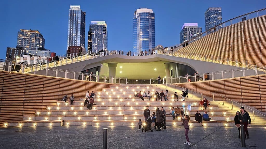 People gather and stroll on the illuminated steps leading to an elevated walkway at the Seattle Waterfront, with skyscrapers glowing in the twilight background.